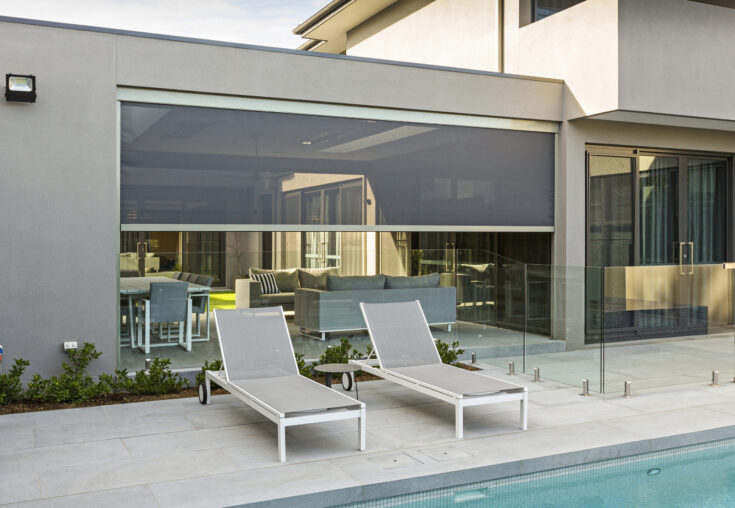 Swimming pool area with two white lounge chairs, glass barrier, and a patio with furniture behind.
